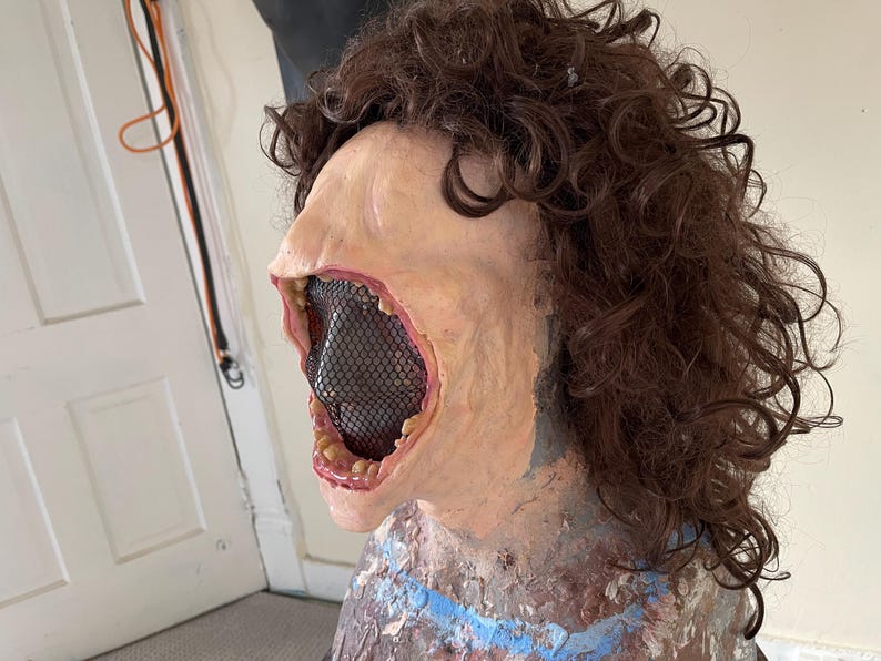 K&ouml;nnte beinhalten: Nahaufnahme einer realistischen Horror-Maske mit weit offenem Mund, die ein schwarzes Netz enth&uuml;llt, das das Innere des Mundes bedeckt. Die Maske hat einen blassen Hautton und ist aus Latex. Die Maske wird auf einem Mannequin mit braunen Locken getragen.