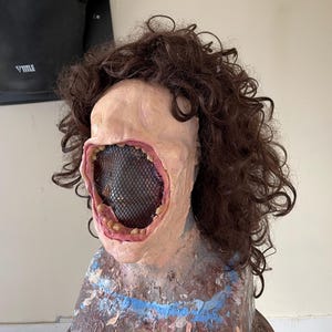 K&ouml;nnte beinhalten: Eine realistisch aussehende Maske mit einem gro&szlig;en, offenen Mund und einem schwarzen Netz, das die Innenseite des Mundes bedeckt. Die Maske ist aus einem fleischfarbenen Material gefertigt und hat braunes, lockiges Haar.