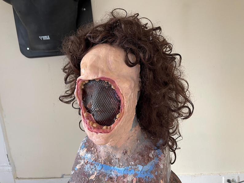 K&ouml;nnte beinhalten: Eine realistisch aussehende Maske mit einem gro&szlig;en, offenen Mund und einem schwarzen Netz, das die Innenseite des Mundes bedeckt. Die Maske ist aus einem fleischfarbenen Material gefertigt und hat braunes, lockiges Haar.