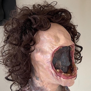 K&ouml;nnte beinhalten: Eine realistische Horror-Maske mit einem gro&szlig;en, offenen Mund, einem schwarzen Netz, das das Innere des Mundes bedeckt, und einer braunen, lockigen Per&uuml;cke.