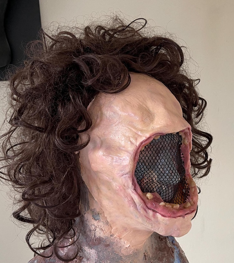 K&ouml;nnte beinhalten: Eine realistische Horror-Maske mit einem gro&szlig;en, offenen Mund, einem schwarzen Netz, das das Innere des Mundes bedeckt, und einer braunen, lockigen Per&uuml;cke.