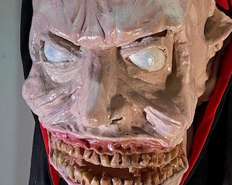 May include: A creepy, pale-skinned mask with a wide, toothy grin. The mask has a red and black hooded robe.