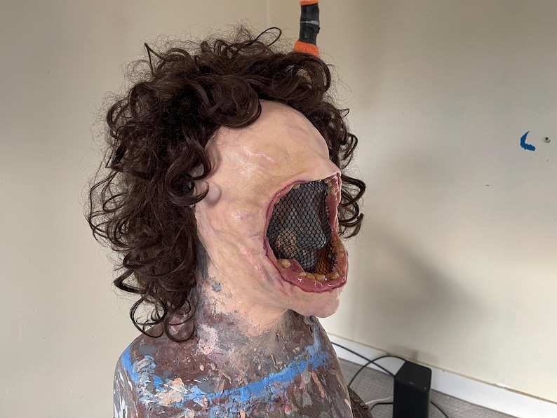 K&ouml;nnte beinhalten: Nahaufnahme einer realistischen Horror-Maske mit einem gro&szlig;en, offenen Mund, einem schwarzen Netz, das das Innere des Mundes bedeckt, und einer braunen, lockigen Per&uuml;cke.