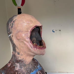 K&ouml;nnte beinhalten: Nahaufnahme einer realistisch aussehenden, blassh&auml;utigen Latexmaske mit einem gro&szlig;en, offenen Mund. Der Mund ist mit einem schwarzen Netz ausgekleidet und hat ein rosafarbenes, fleischiges Inneres. Die Maske befindet sich auf einem Mannequin-Kopf.