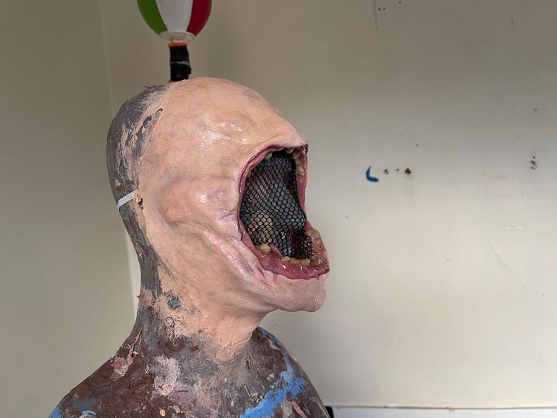 K&ouml;nnte beinhalten: Nahaufnahme einer realistisch aussehenden, blassh&auml;utigen Latexmaske mit einem gro&szlig;en, offenen Mund. Der Mund ist mit einem schwarzen Netz ausgekleidet und hat ein rosafarbenes, fleischiges Inneres. Die Maske befindet sich auf einem Mannequin-Kopf.