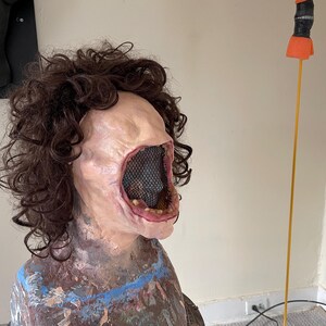 K&ouml;nnte beinhalten: Eine realistisch aussehende, blasse Maske mit einem gro&szlig;en, offenen Mund und einem schwarzen Netz, das das Innere des Mundes bedeckt. Die Maske hat eine braune, lockige Per&uuml;cke.
