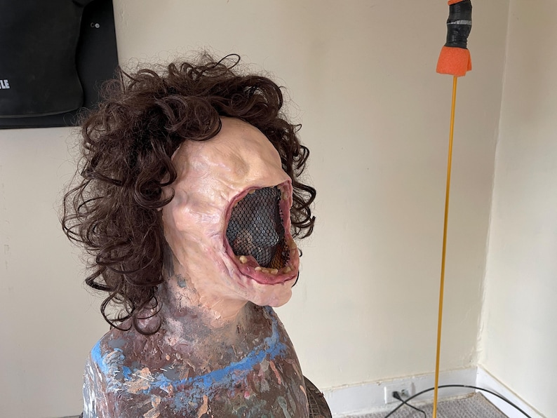 K&ouml;nnte beinhalten: Eine realistisch aussehende, blasse Maske mit einem gro&szlig;en, offenen Mund und einem schwarzen Netz, das das Innere des Mundes bedeckt. Die Maske hat eine braune, lockige Per&uuml;cke.