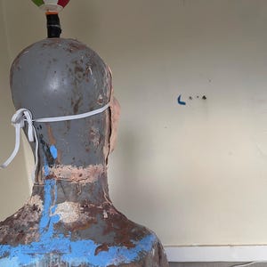 K&ouml;nnte beinhalten: Ein grauer Mannequin-Kopf mit einer wei&szlig;en Gesichtsmaske. Der Mannequin ist mit blauer, brauner und wei&szlig;er Farbe bemalt. Der Mannequin ist von der Kamera abgewandt.