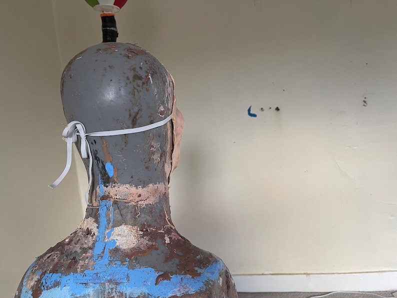 K&ouml;nnte beinhalten: Ein grauer Mannequin-Kopf mit einer wei&szlig;en Gesichtsmaske. Der Mannequin ist mit blauer, brauner und wei&szlig;er Farbe bemalt. Der Mannequin ist von der Kamera abgewandt.