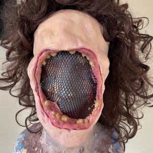 K&ouml;nnte beinhalten: Nahaufnahme einer realistischen Horror-Maske mit einem gro&szlig;en, offenen Mund und einem schwarzen Netz, das das Innere des Mundes bedeckt. Die Maske ist aus einem fleischfarbenen Material gefertigt und hat eine realistische Textur.