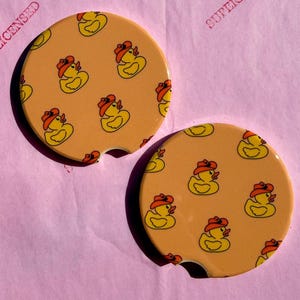 May include: Two round car coasters with a yellow background and a repeating pattern of yellow cartoon ducks wearing red cowboy hats. The coasters have a small notch for easy removal and are on a pink surface.