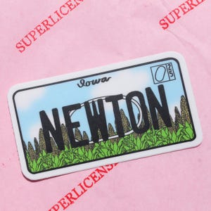 Puede incluir: Una pegatina rectangular que se asemeja a una placa de matrícula de Iowa, con la palabra "NEWTON" en letras negras en negrita. El diseño incluye un fondo de cielo azul, mazorcas de maíz y la palabra "Iowa" en la parte superior. La pegatina está sobre un fondo rosa.