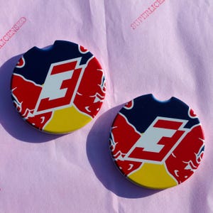 May include: Two round car coasters with a navy blue, red, white, and yellow design. The coasters feature a white stylized letter "E" and a red bull logo. The coasters have a small notch for easy removal from the car cup holder.