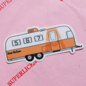 May include: A sticker of a retro camper in orange and white, with the numbers 5, 6, and 7 displayed. The text "Arrow McLaren" is printed on the side. The sticker is on a pink background with the word "SUPERLICEN" in red.