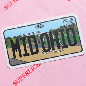Puede incluir: Una pegatina rectangular con borde blanco que presenta el diseño de una matrícula. La matrícula dice "MID OHIO" en letras negras grandes, con "Ohio" encima. El fondo muestra una escena de campo de golf con árboles y cielo azul.