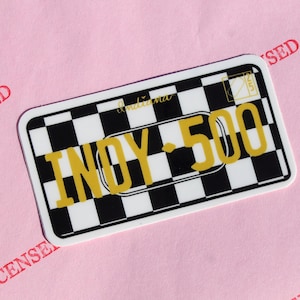 Puede incluir: Una pegatina rectangular con un patrón de cuadros blancos y negros, con el texto "INDY 500" en amarillo. La palabra "Indiana" está escrita en cursiva en la parte superior. La pegatina tiene un borde blanco y está sobre un fondo rosa.