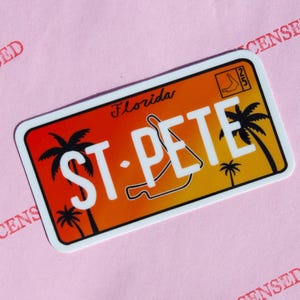 Puede incluir: Adhesivo rectangular que simula una placa de matrícula de Florida, con "ST. PETE" en letras blancas. El fondo transiciona de rojo a amarillo, con siluetas de palmeras. La palabra "Florida" está arriba.