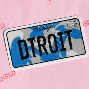 Puede incluir: Una pegatina rectangular que se asemeja a una placa de matrícula de Michigan, con el nombre de la ciudad "DETROIT" en letras negras en negrita. El fondo tiene un patrón de camuflaje azul y gris, con "Michigan" en cursiva.