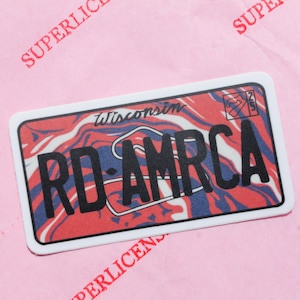 Puede incluir: Una pegatina rectangular que se asemeja a una placa de matrícula de Wisconsin. El diseño presenta un patrón veteado rojo, blanco y azul con el texto "RD-AMRCA" en letras negras en negrita. La palabra "Wisconsin" está arqueada sobre el centro de la placa.