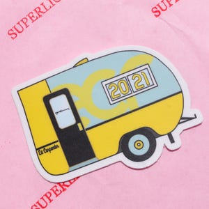 May include: A sticker featuring a yellow and light blue camper with a black door and a wheel. The camper has the year "2021" displayed on the side. The sticker has the text "Ed Carpenter" on the side.