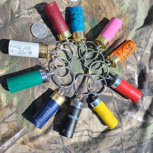 May include: A collection of ten shotgun shell keychains in various colors, including red, blue, green, yellow, pink, and black. Each keychain features a different brand name and details, such as "Featherlite 1150 FPS" and "Clever Mirage".