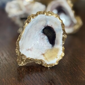 May include: Close-up of a decorative oyster shell with a gold-painted edge. The shell interior is white with a dark, blackish-purple area. The shell is on a dark wooden surface.