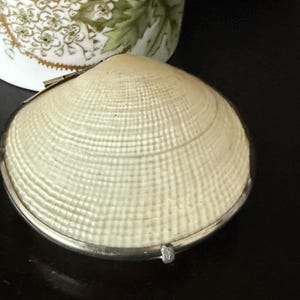 May include: A small, round, off-white clam shell box with a textured, grid-like surface pattern. The shell is encased in a thin, polished silver-toned metal rim with a small hinge and a clasp mechanism visible at the front edge of the container.