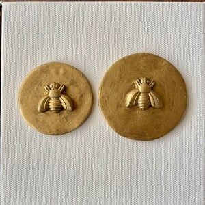 May include: Two gold-colored round wall decorations with a raised bee design. The bee is in the center of each circle and has a detailed design.