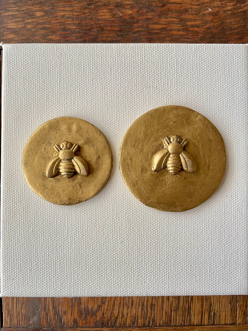 May include: Two gold-colored round wall decorations with a raised bee design. The bee is in the center of each circle and has a detailed design.