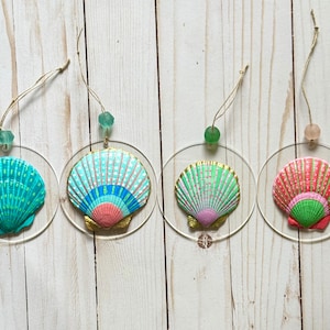 Puede incluir: Cuatro adornos de conchas marinas con respaldos circulares transparentes. Cada concha está pintada en colores vibrantes: turquesa, azul, rosa y verde. Cuelgan de hilo con una cuenta en la parte superior, ideal para decoración costera.
