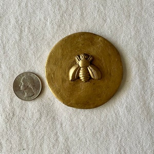May include: A gold-colored round coaster with a raised bee design. The coaster is about the size of a quarter.