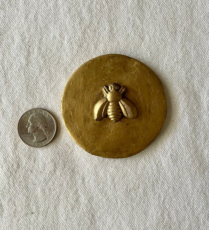 May include: A gold-colored round coaster with a raised bee design. The coaster is about the size of a quarter.
