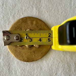 May include: A gold-colored metal disc with a raised design, measured with a tape measure to be approximately 2.5 inches in diameter.