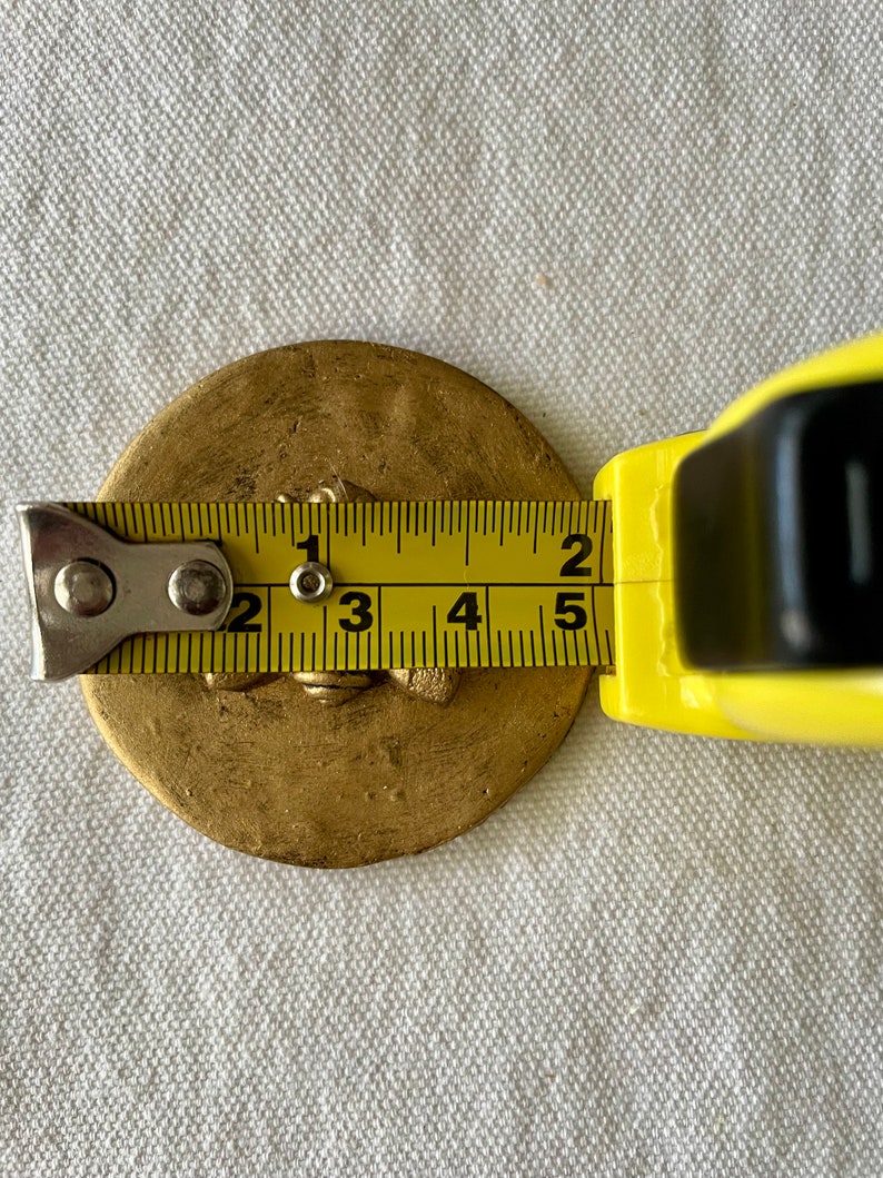 May include: A gold-colored metal disc with a raised design, measured with a tape measure to be approximately 2.5 inches in diameter.
