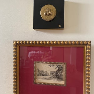 May include: A framed black and white etching of a building with trees on either side. The etching is framed in a gold frame with a red mat. A small gold bee sculpture is mounted on a black square.