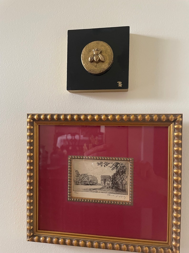 May include: A framed black and white etching of a building with trees on either side. The etching is framed in a gold frame with a red mat. A small gold bee sculpture is mounted on a black square.