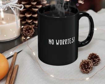 May include: Black coffee mug with the text "NO WORRIES!" in white lettering. The mug is sitting on a white surface with string lights and pine cones.