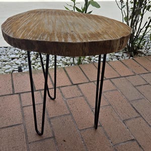 May include: A round wooden side table with a natural wood finish and black hairpin legs. The table is sitting on a brick patio.