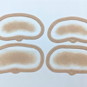 Pode incluir: Quatro stencils de maquiagem bege com uma borda ligeiramente elevada. Cada stencil tem um remendo marrom e felpudo no centro e a letra "M" impressa na parte inferior.