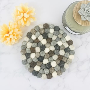 May include: A round gray, brown, and white felt coaster made of small balls. The coaster is on a white marble surface.