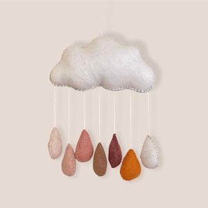 May include: A felt cloud mobile with seven felt raindrops hanging from it. The cloud is white and the raindrops are in shades of beige, pink, brown, and orange.