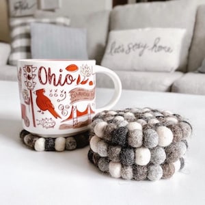 May include: A white ceramic mug with a red and black illustration of Ohio landmarks and the text "Ohio". The mug is sitting on a round, gray and white felt coaster.