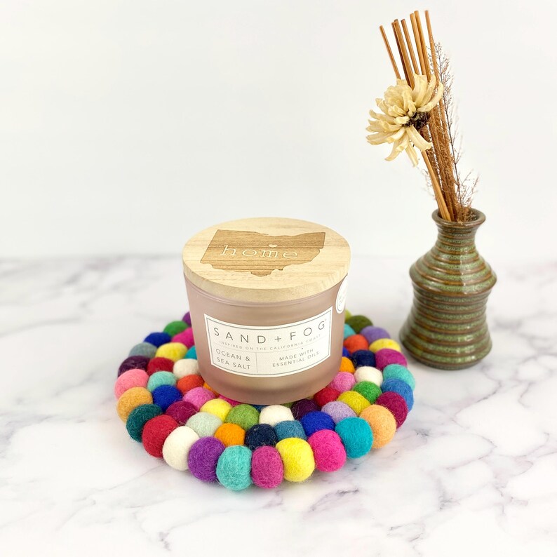 K&ouml;nnte beinhalten: Eine mattgl&auml;nzende Kerze aus Glas mit einem Holzdeckel, auf dem "home" steht. Die Kerze steht auf einem bunten Filzuntersetzer und ist mit "Sand + Fog" und "Ocean & Sea Salt" beschriftet.