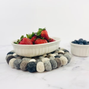 K&ouml;nnte beinhalten: Eine wei&szlig;e ovale Keramikschale gef&uuml;llt mit frischen roten Erdbeeren steht auf einem grauen und wei&szlig;en Filzuntersetzer. Der Untersetzer besteht aus kleinen, runden Filzkugeln.