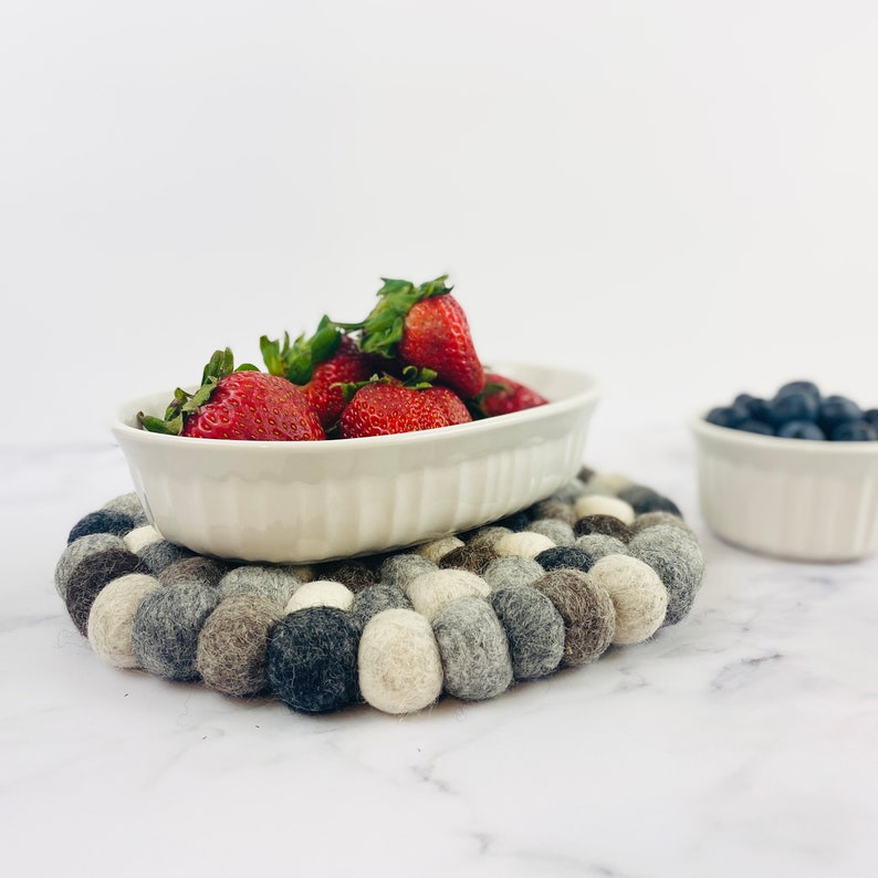K&ouml;nnte beinhalten: Eine wei&szlig;e ovale Keramikschale gef&uuml;llt mit frischen roten Erdbeeren steht auf einem grauen und wei&szlig;en Filzuntersetzer. Der Untersetzer besteht aus kleinen, runden Filzkugeln.