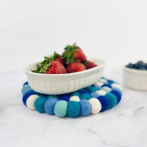 K&ouml;nnte beinhalten: Eine wei&szlig;e ovale Keramikschale gef&uuml;llt mit frischen Erdbeeren steht auf einem blauen und wei&szlig;en Filzuntersetzer. Der Untersetzer besteht aus kleinen, runden Filzkugeln.