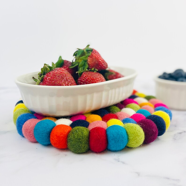 K&ouml;nnte beinhalten: Eine wei&szlig;e Keramik-Backform gef&uuml;llt mit frischen Erdbeeren steht auf einem bunten Filzuntersetzer aus runden Kugeln. Der Untersetzer ist ein lebendiges Gemisch aus Rot, Rosa, Gelb, Gr&uuml;n, Blau und Lila.