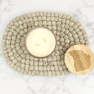 May include: A white candle with two wicks sits on a gray felt coaster. The candle is in a clear glass jar with a light pink rim. A wooden coaster with the word "home" engraved on it is next to the candle.