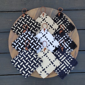 May include: Nine black and white woven pot holders with brown leather loops. The pot holders are arranged in a circle on a wooden surface.