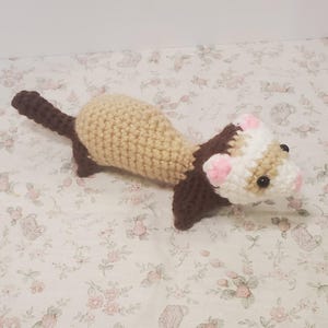 May include: A handmade crochet ferret toy. The ferret has a tan body, a brown tail and legs, and a head with brown, white, and pink details. The ferret has black safety eyes. The toy is made of soft yarn.